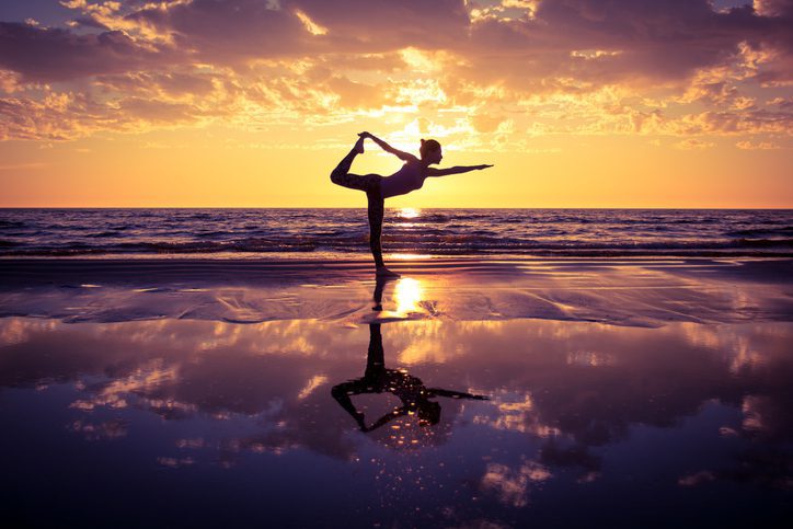 Lady in yoga pose, on the sand at the beach in front of a sunset