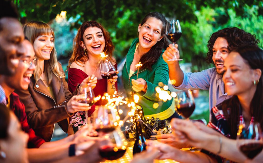 Happy friends having fun with fire sparkles while toasting red wine - Young millenial people camping at summer picnic barbecue party on night mood - Youth life style concept on warm vivid filter